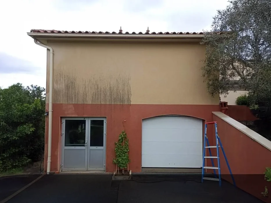 Scène extérieure d'une terrasse pavée en pierre naturelle devant une maison traditionnelle avec volets blancs et végétation grimpante